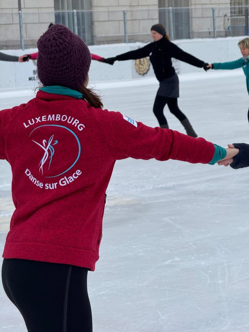 Patineurs se tenant par la main sur une patinoire extérieure, avec une veste rouge Luxembourg Danse sur Glace au premier plan